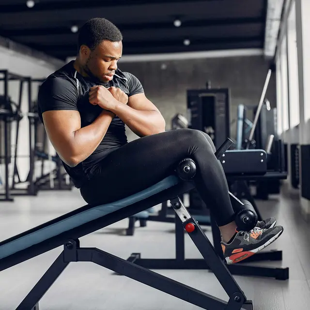 A man performing a seated exercise in a gym, wearing a black workout outfit and focused on his form. The gym is equipped with various fitness machines in the background.
