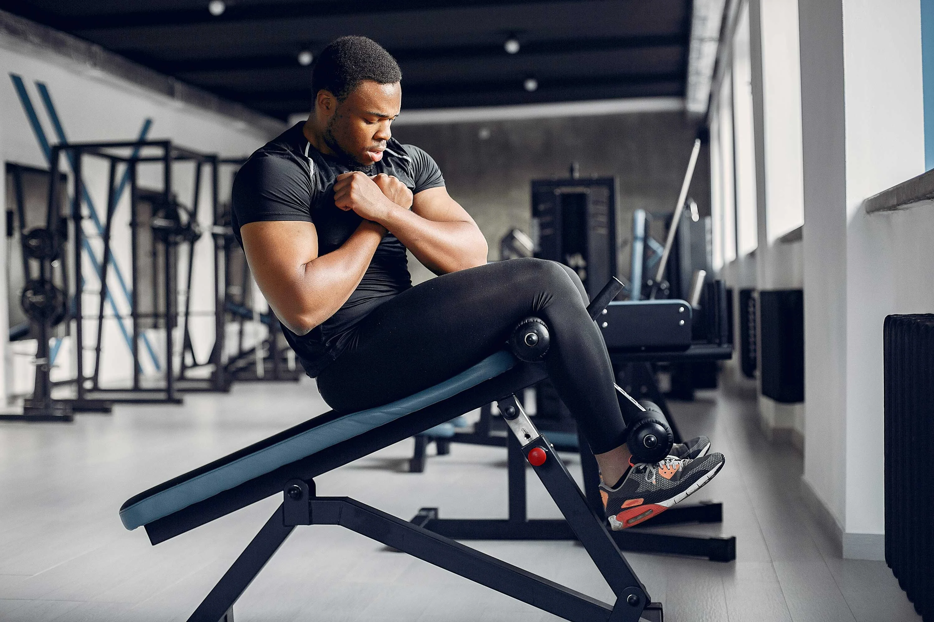 A man performing a seated exercise in a gym, wearing a black workout outfit and focused on his form. The gym is equipped with various fitness machines in the background.