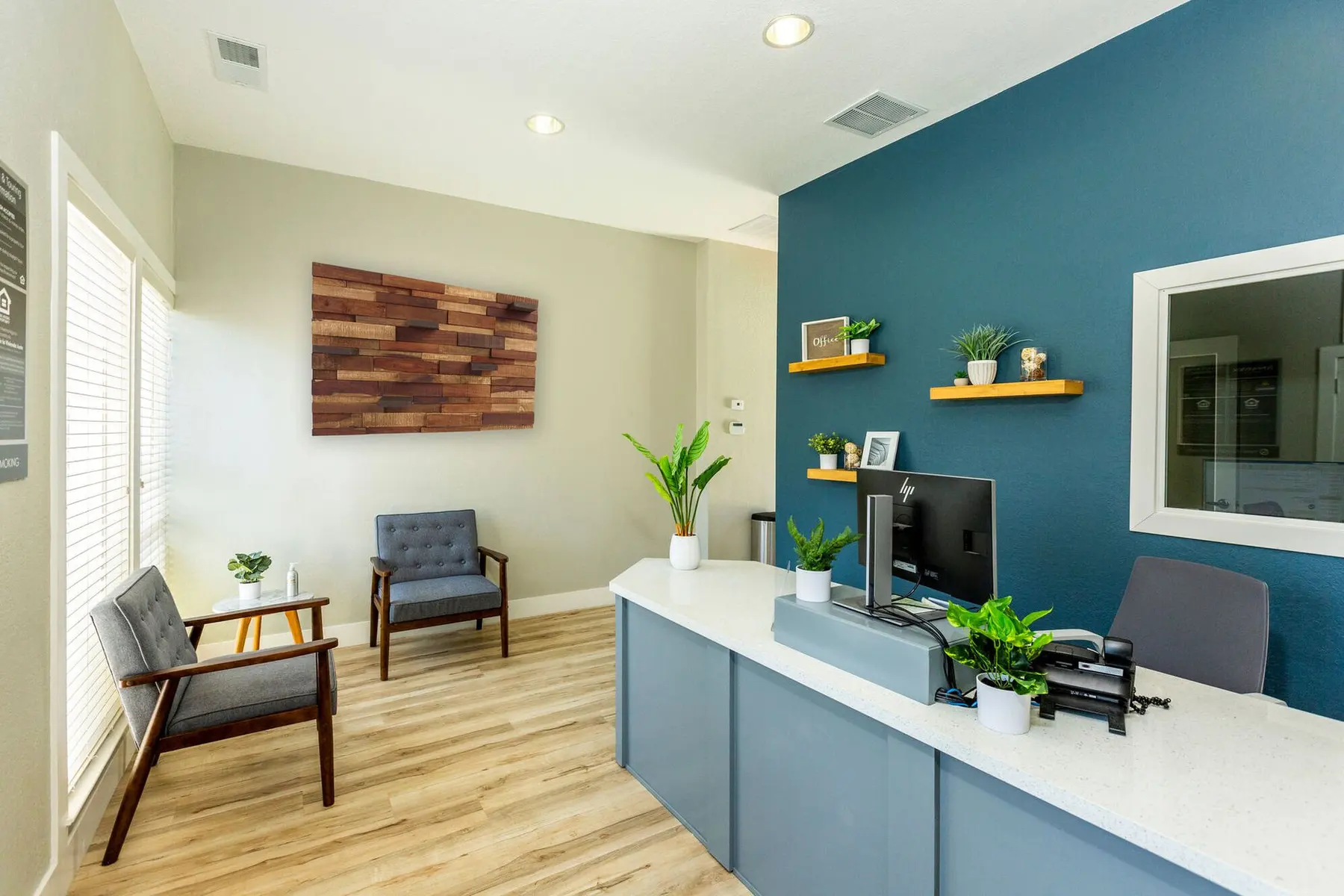 A modern office reception area featuring a blue accent wall, a desk with computer and plants, two chairs, and decorative wall art.