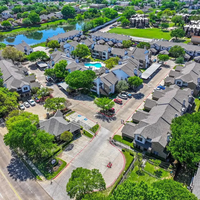Aerial view of a residential complex with several buildings, surrounded by lush greenery, a swimming pool, and a nearby pond.