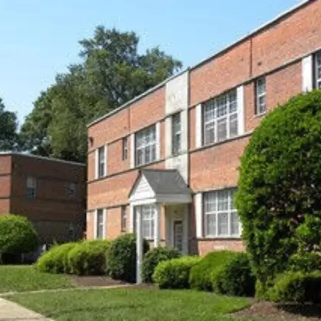 A brick apartment building with landscaped greenery in front, featuring neatly trimmed bushes and a well-maintained lawn under a clear blue sky.
