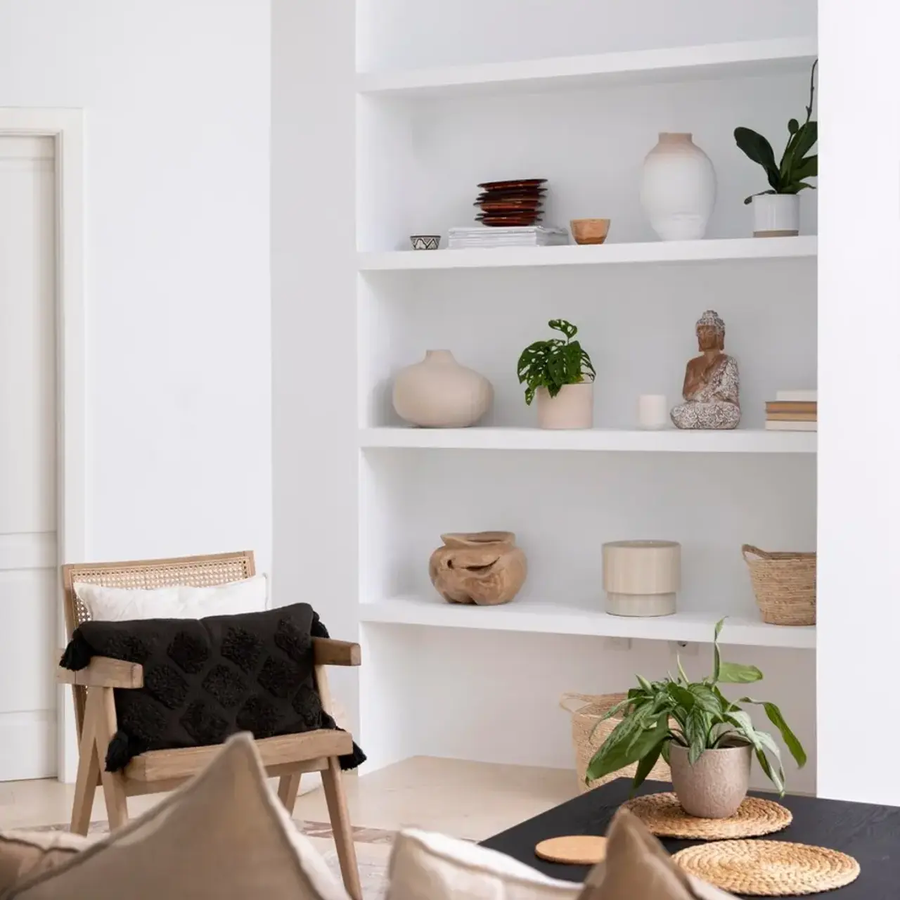 A minimalist living room featuring a beige couch with decorative pillows, a sustainable wooden chair with a throw blanket, and a white shelving unit displaying various decorative items and plants.