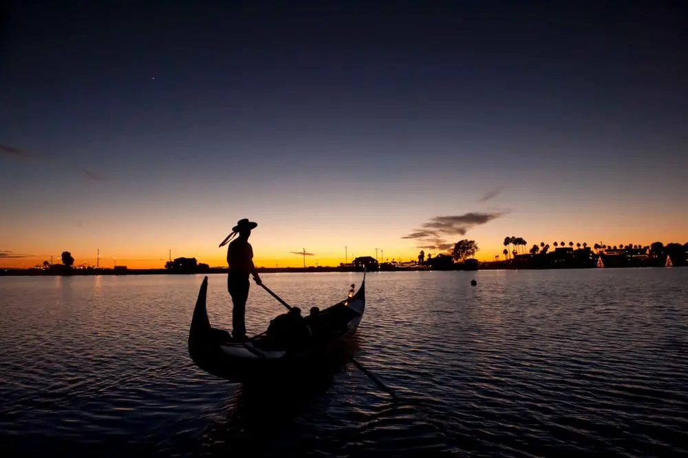 Gondola at Sunset A silhouette of a gondola on calm waters at sunset, with a person standing at the front and two people seated in the gondola.