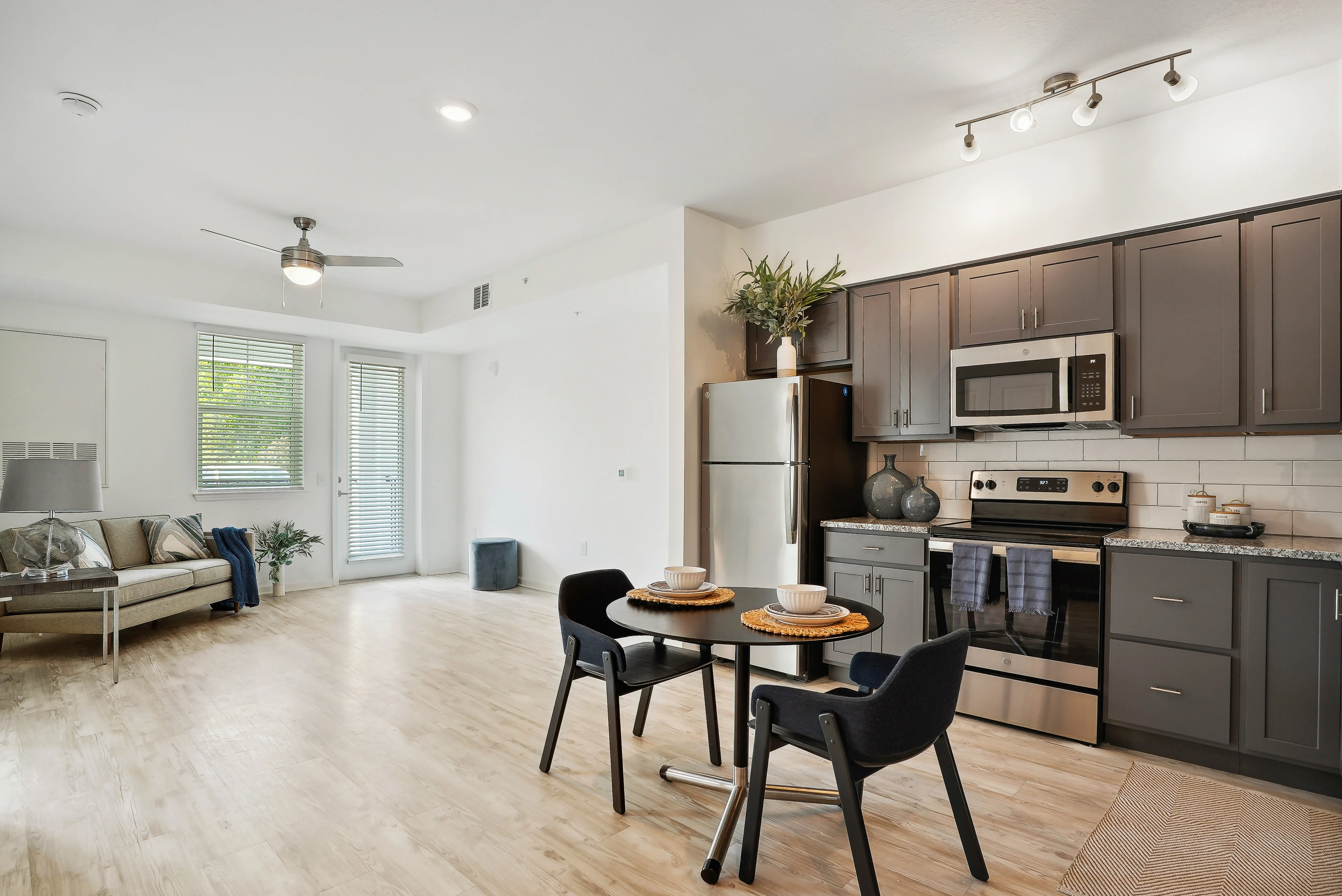 Contemporary Apartment Living Space A modern apartment interior featuring a kitchen, dining area, and living space. The kitchen has dark cabinets and stainless steel appliances. A small dining table is set for two. Natural light fills the room from large windows and glass doors, leading to an outdoor space.