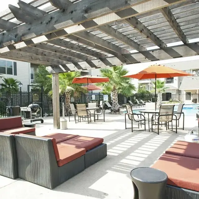 A spacious outdoor lounge area next to a pool, featuring a pergola with shaded seating, tables, and umbrellas.