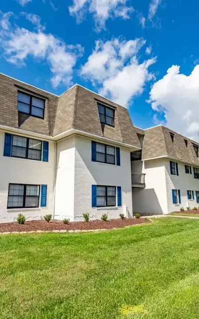 Exterior view of a multi-story apartment building with blue window shutters, surrounded by green grass and trees under a cloudy sky.