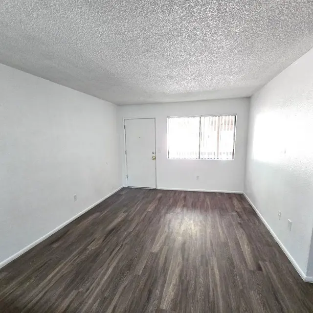 Vacant Room Interior A vacant room with a textured ceiling, white walls, and wooden flooring. There is a door on the left side and a window with blinds on the opposite wall, letting in natural light. A small dark doorway is visible on the right leading to another room.