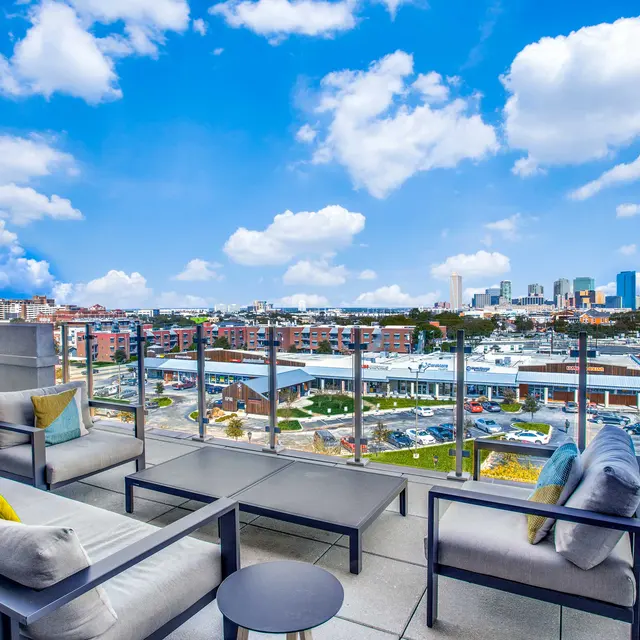 A rooftop lounge area with modern furniture overlooking a city skyline on a bright, sunny day with a partly cloudy sky.