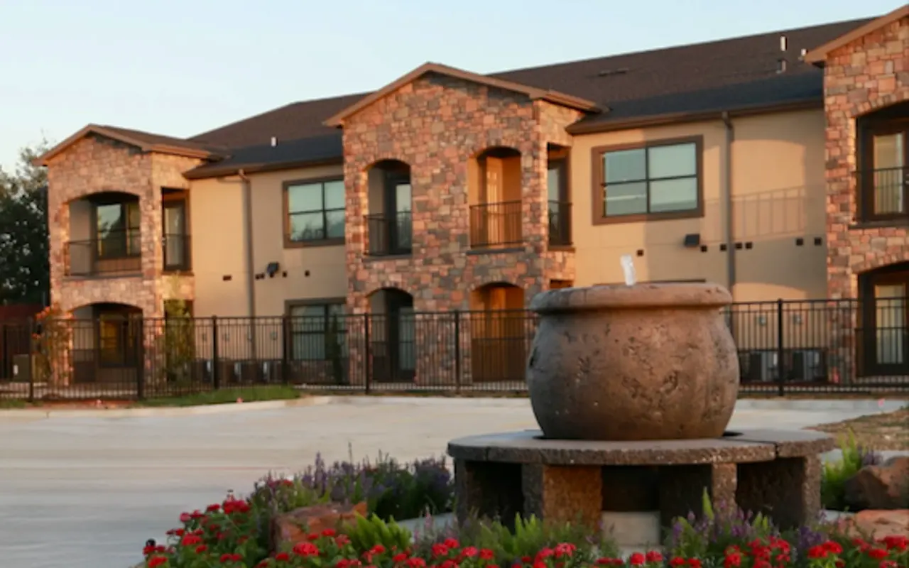 A modern apartment complex featuring stone exterior and balcony, surrounded by landscaped flowerbeds with red flowers and a decorative fountain at the foreground.
