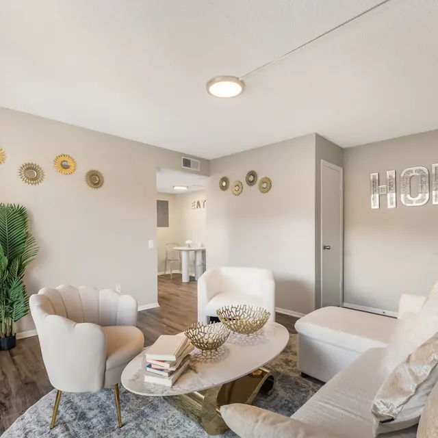 A modern living room featuring two white chairs, a round coffee table with decor, and green plants. The walls are decorated with circular wall art and the word 'HOME' in shiny letters.