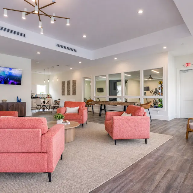 A modern lounge area featuring four pink armchairs arranged around a small table on a large beige rug. The space is well-lit with recessed lighting and a central chandelier. In the background, there is a large mirror wall, a wooden table, and a television on the wall displaying a colorful image. A potted plant adds a touch of greenery to the room.