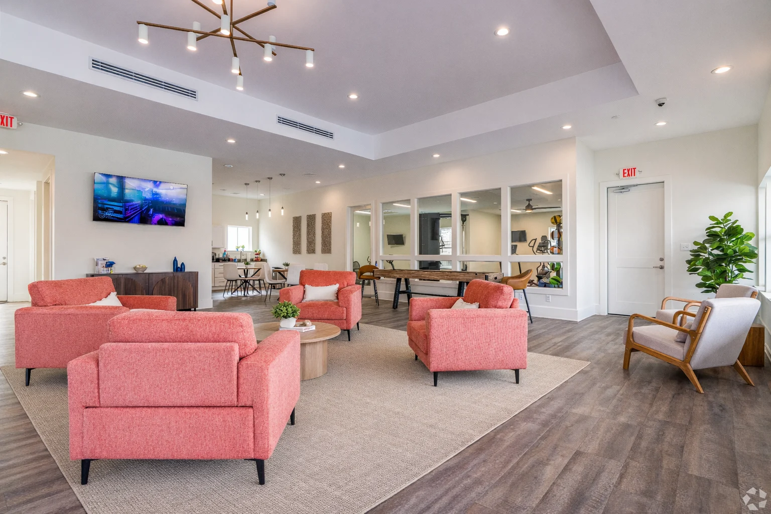 A modern lounge area featuring four pink armchairs arranged around a small table on a large beige rug. The space is well-lit with recessed lighting and a central chandelier. In the background, there is a large mirror wall, a wooden table, and a television on the wall displaying a colorful image. A potted plant adds a touch of greenery to the room.