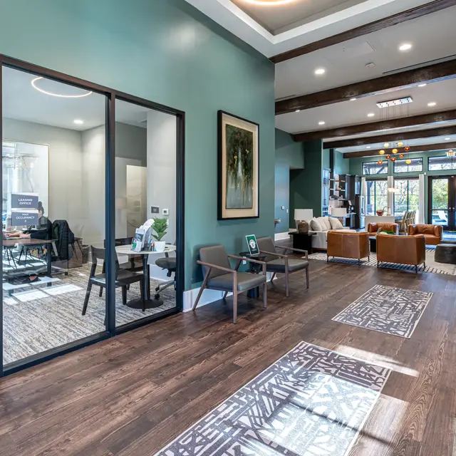 A spacious modern lobby with a divided seating area and a glass-walled meeting room, featuring wooden beams, stylish furniture, and decorative rugs.