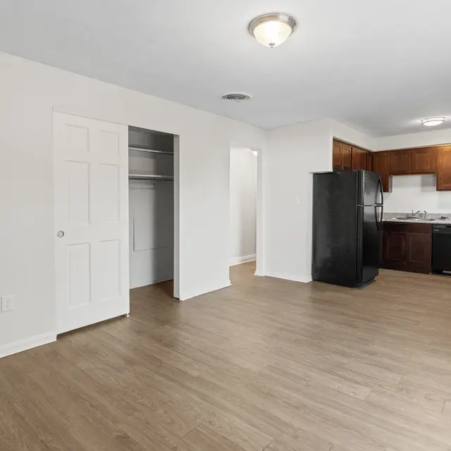 Interior view of a modern apartment featuring an open floor plan with a kitchen and living area. The kitchen has dark wooden cabinets and black appliances. The flooring is light-colored laminate.
