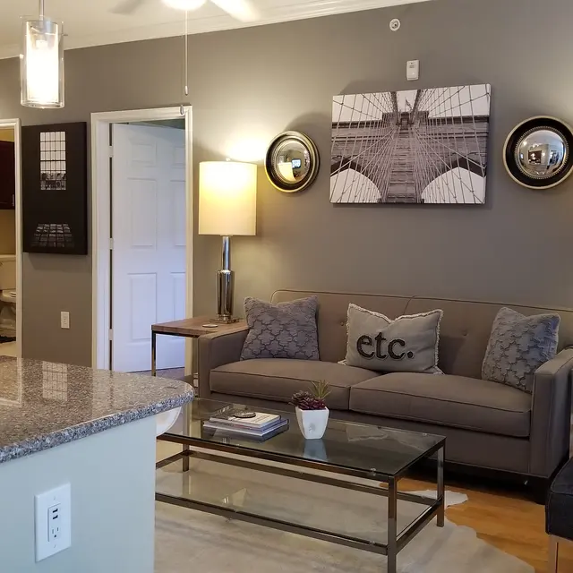 A modern living room featuring a beige couch with decorative pillows, a glass coffee table, and two lamps. The walls are painted gray with framed artwork, and a kitchen area with a bar counter is visible in the foreground.