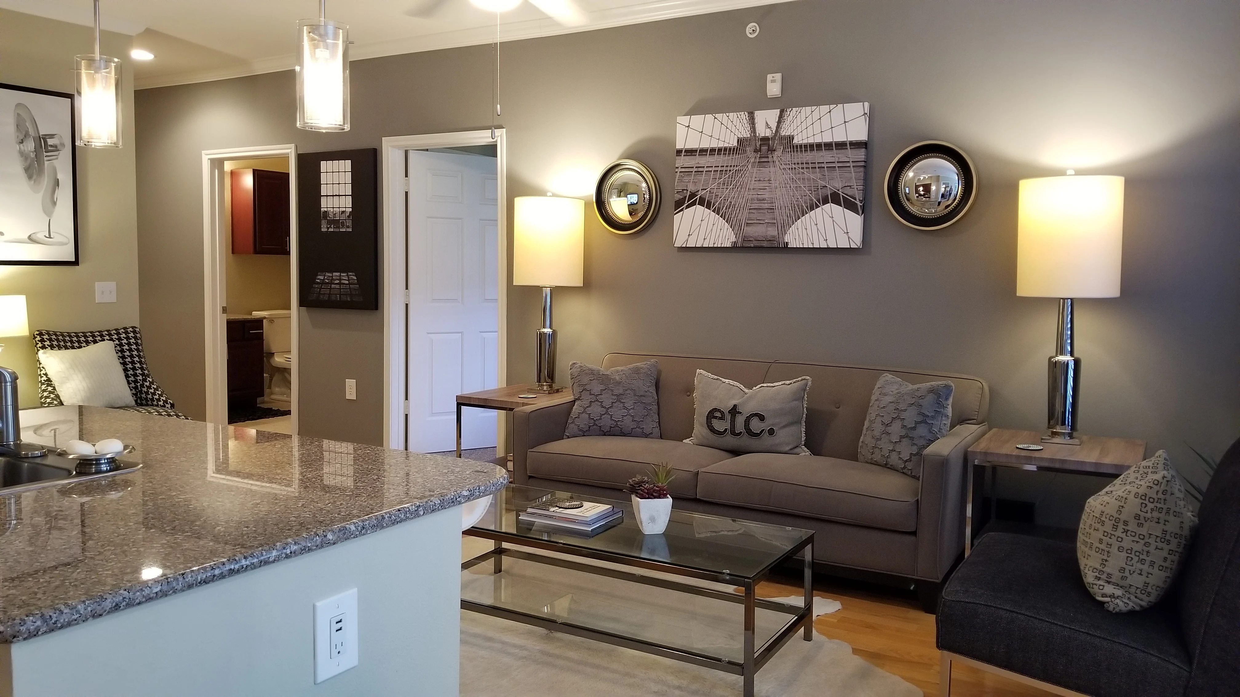 Stylish Modern Living Room A modern living room featuring a beige couch with decorative pillows, a glass coffee table, and two lamps. The walls are painted gray with framed artwork, and a kitchen area with a bar counter is visible in the foreground.