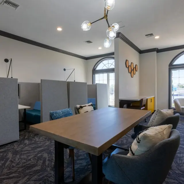 A modern workspace featuring a large table surrounded by comfortable chairs, semi-private booths on the left, and large windows letting in natural light.