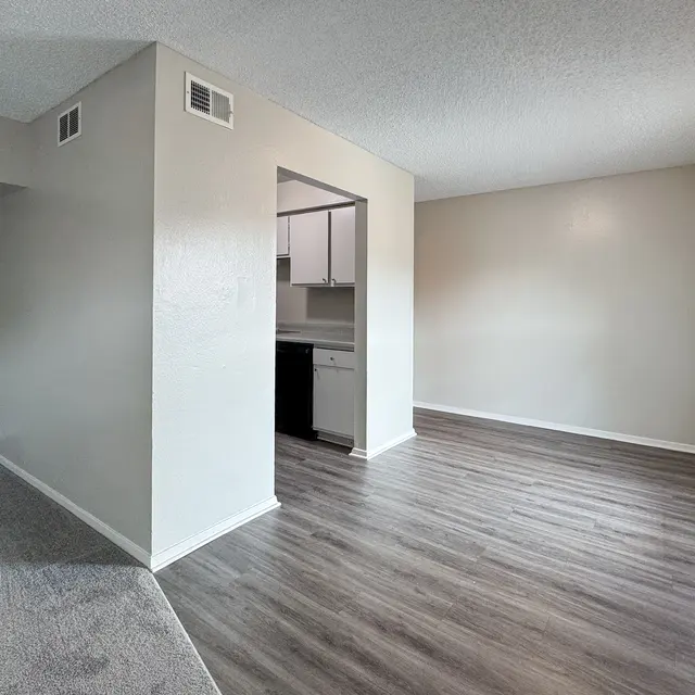 A modern apartment interior featuring a spacious living area with new flooring, a partially visible kitchen, and ample natural light from a window.