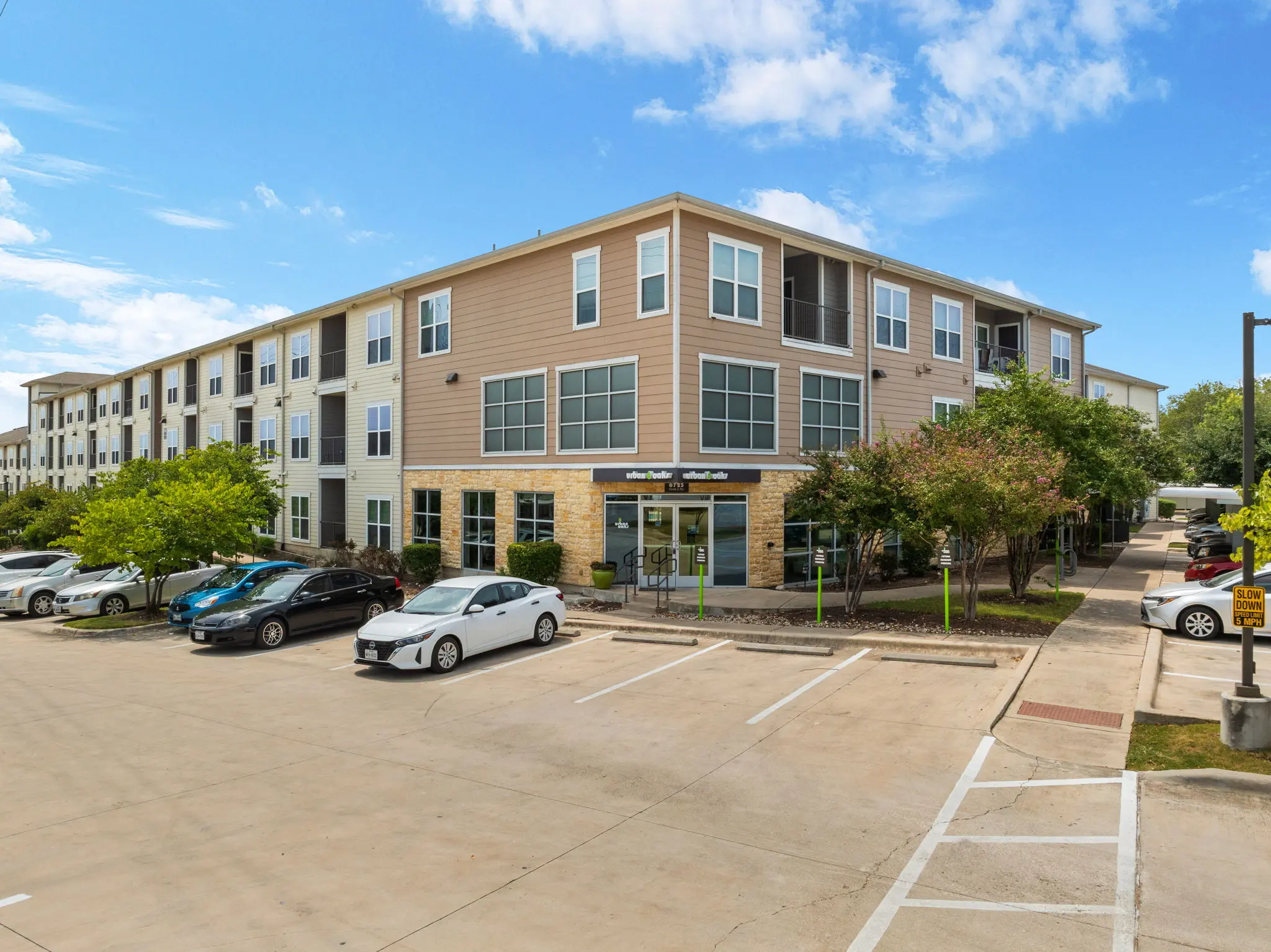 A modern apartment complex with multiple stories and a landscaped parking area. Cars are parked in front and trees provide greenery around the building under a clear blue sky.