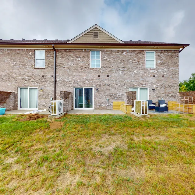A view of the back of a two-story brick apartment building with several patio areas. The grassy area in front is mostly dry and beige, and there are a few patio furniture pieces, including a couple of chairs and tables. Air conditioning units are visible under the windows, and the sky is partly cloudy.