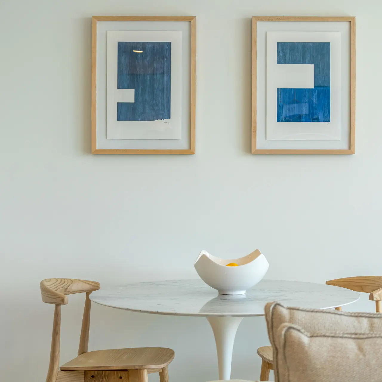 A modern interior featuring a round marble table with wooden chairs, a decorative bowl in the center, and two framed blue abstract art pieces on the wall.