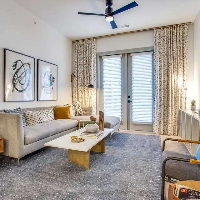A stylish modern living room featuring a gray sofa with patterned throw pillows, a light wood coffee table adorned with decor, a blue accent chair, and a media console with a turntable. Large windows with sheer curtains let in natural light, creating a welcoming atmosphere.
