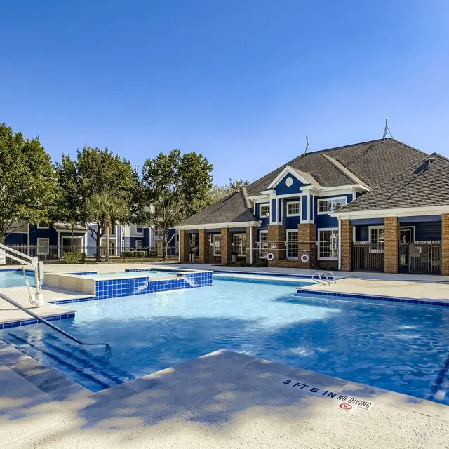 A spacious swimming pool area featuring a large pool with a shallow section, surrounded by sun loungers and lush greenery. The clubhouse can be seen in the background with a charming architectural design.