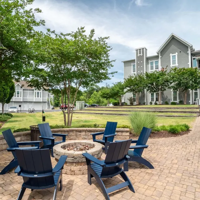 Outdoor seating area with blue chairs arranged around a stone fire pit, surrounded by greenery and buildings.