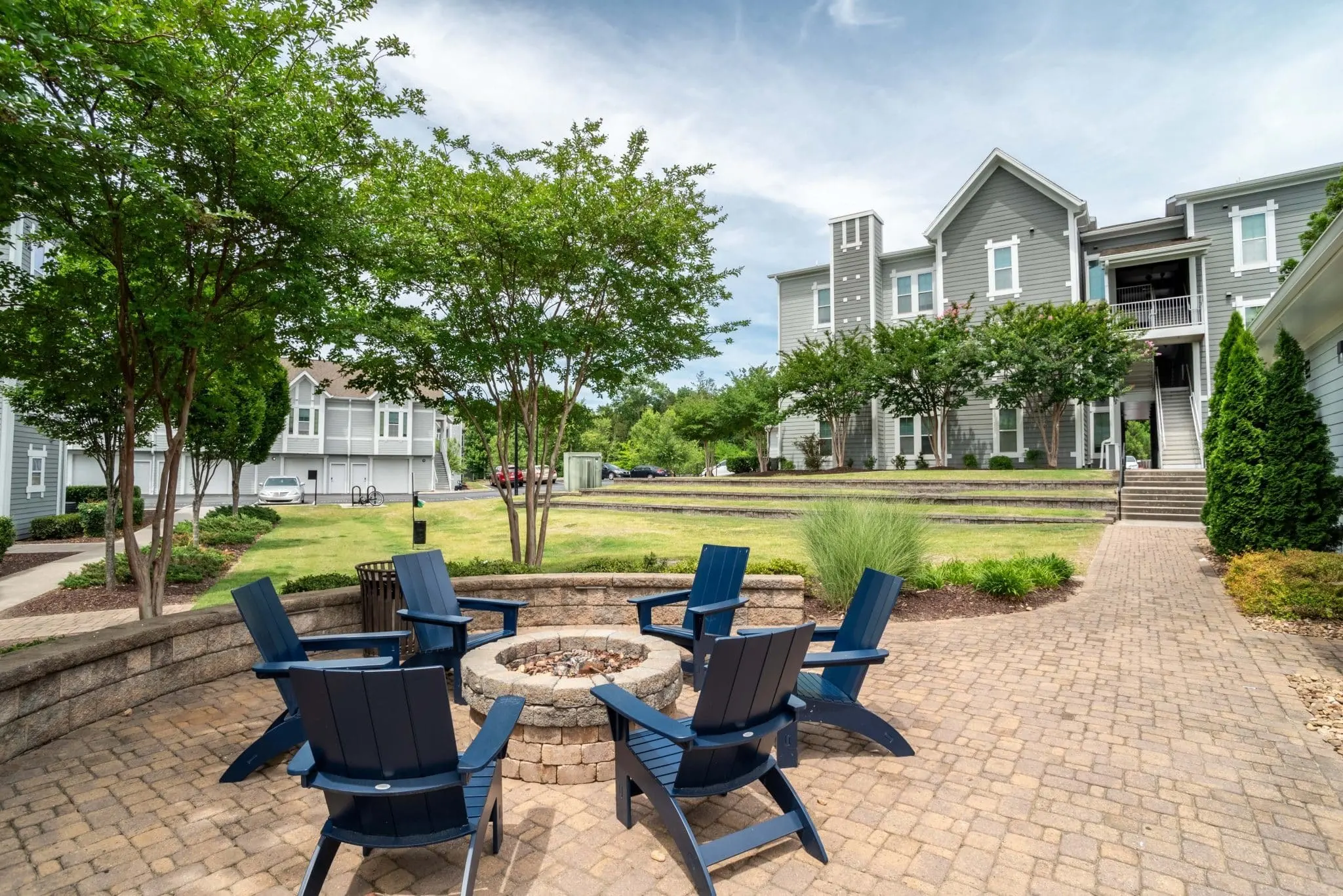 Outdoor Gathering Space Outdoor seating area with blue chairs arranged around a stone fire pit, surrounded by greenery and buildings.