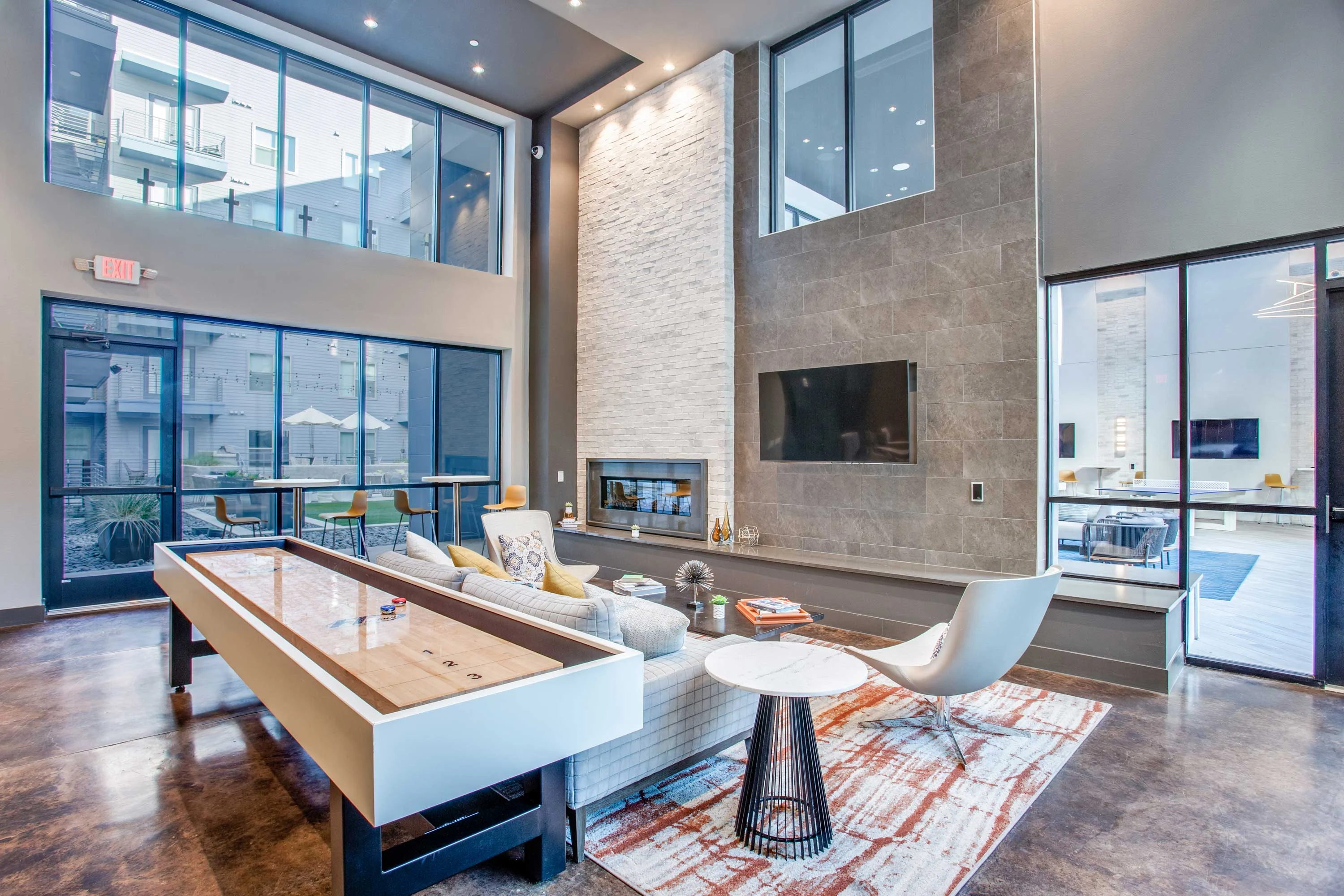 A modern lounge area featuring a stylish fireplace, large windows, and a shuffleboard table, with contemporary furniture and decor.