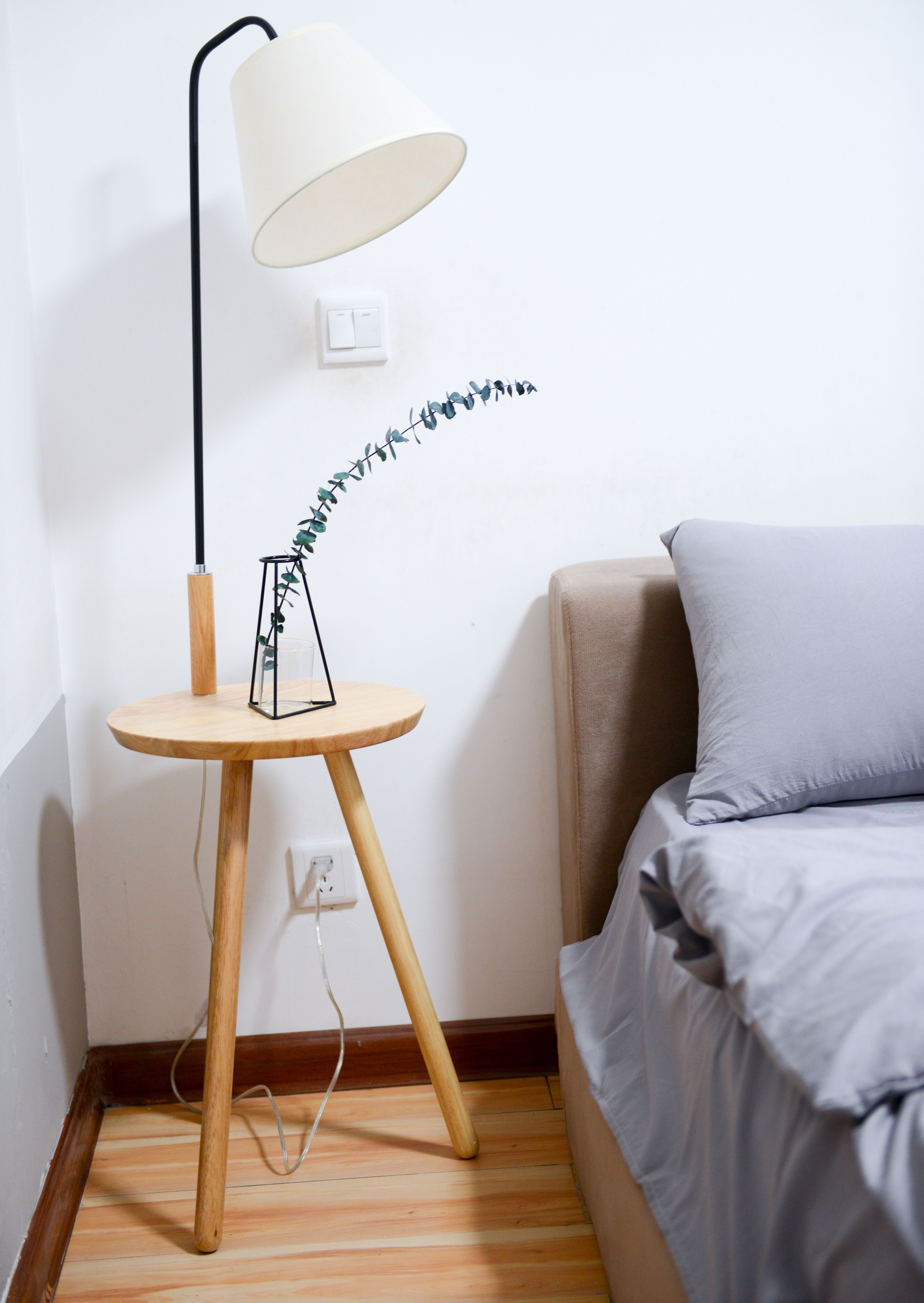 A wooden side table with a modern lamp and a plant vase next to a gray bed in a minimalistic bedroom.