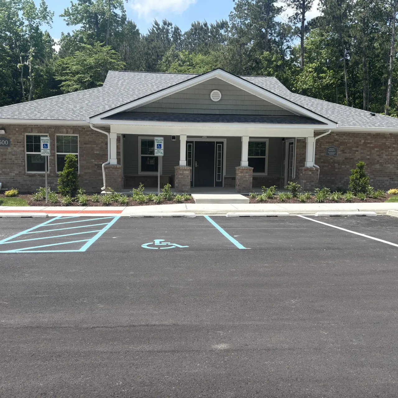 A newly constructed single-story building with a well-designed facade. The structure features a peaked roof, stone accents, and multiple windows. It is surrounded by a landscaped area and has a parking lot with marked spaces, including a wheelchair accessible spot.
