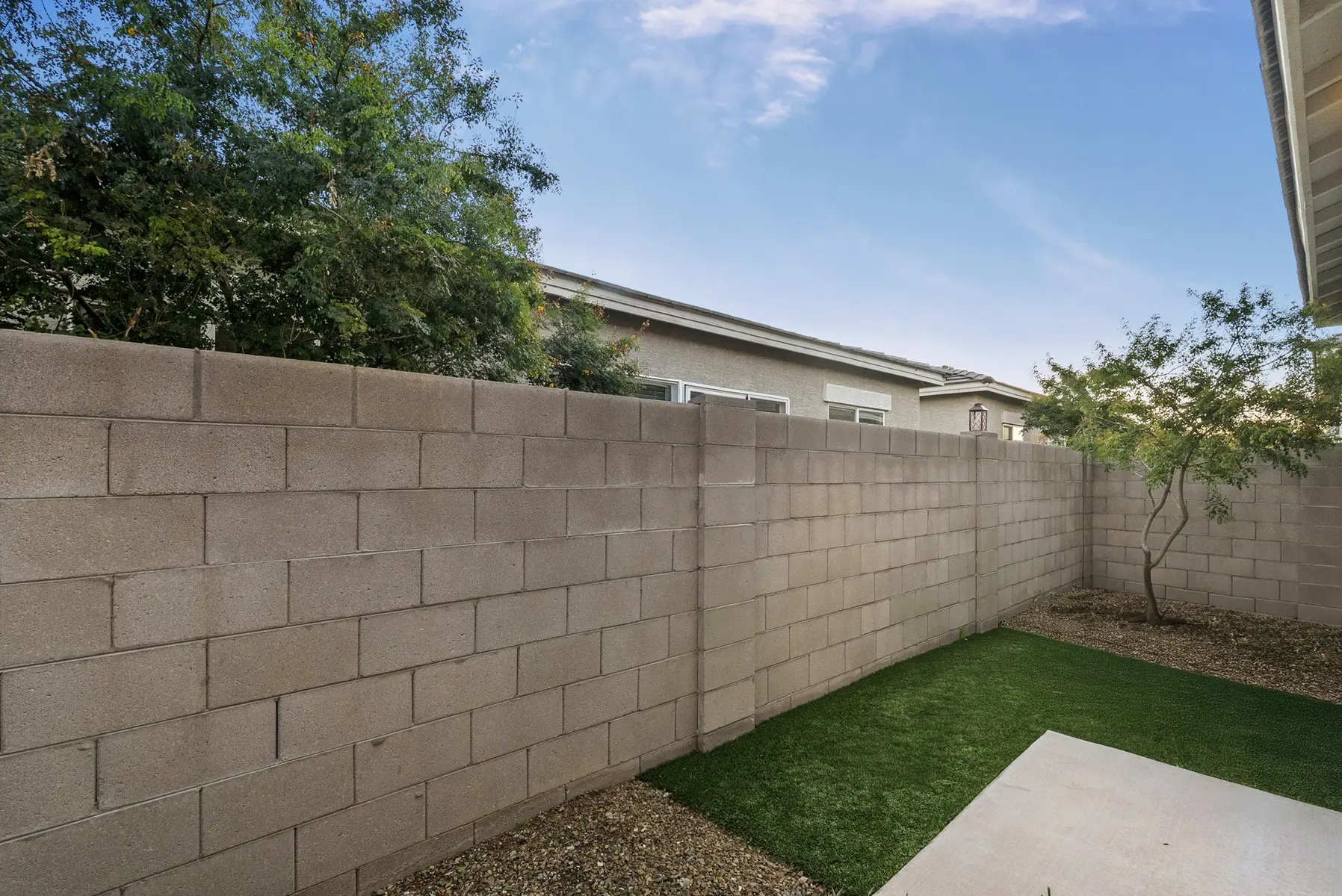 A backyard with a cinder block wall, small grassy area, gravel, and a tree; residential homes in the background under a clear blue sky.