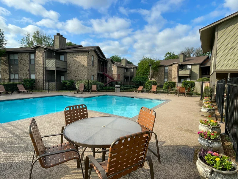 A clear swimming pool surrounded by lounge chairs, with apartment buildings and potted flowers in the background under a blue sky.
