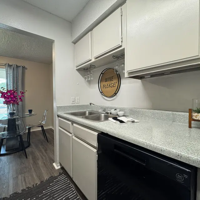 A modern kitchen featuring gray countertops, white cabinets, and a black dishwasher. In the background, there's a dining area with a round glass table and chairs, accompanied by a window with gray curtains. A decorative sign saying 'WRITE PLEASE!' hangs on the wall.