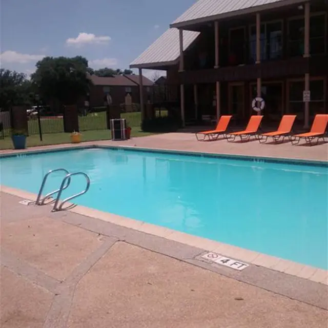 A clear blue swimming pool with a ladder, surrounded by orange lounge chairs under a sunny sky. A building with a gray roof is visible in the background along with a grassy area.