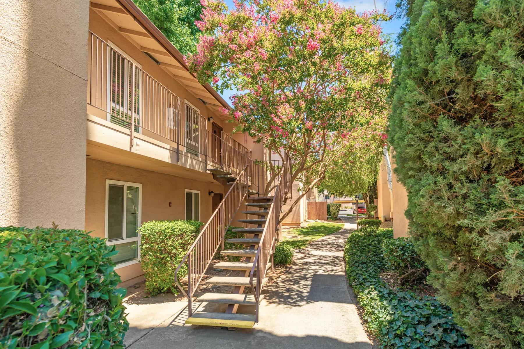 A walkway in an apartment complex with stairs leading to upper floors, surrounded by greenery and colorful trees.