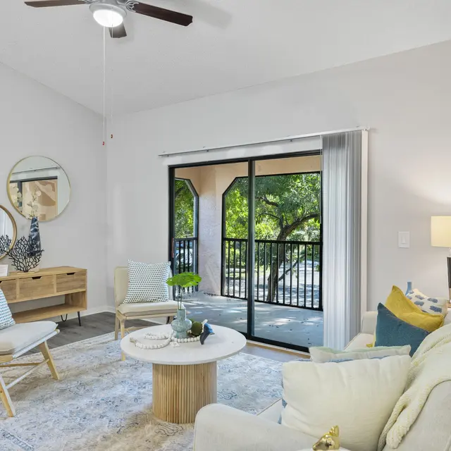 A modern living room with light-colored walls, a round coffee table, beige armchairs, a comfortable sofa adorned with cushions, and a view of a green outdoor area through glass doors.
