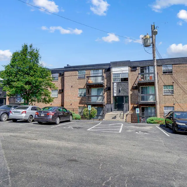 View of a multi-story apartment building with a parking lot in front, featuring several parked cars and trees nearby under a clear blue sky.
