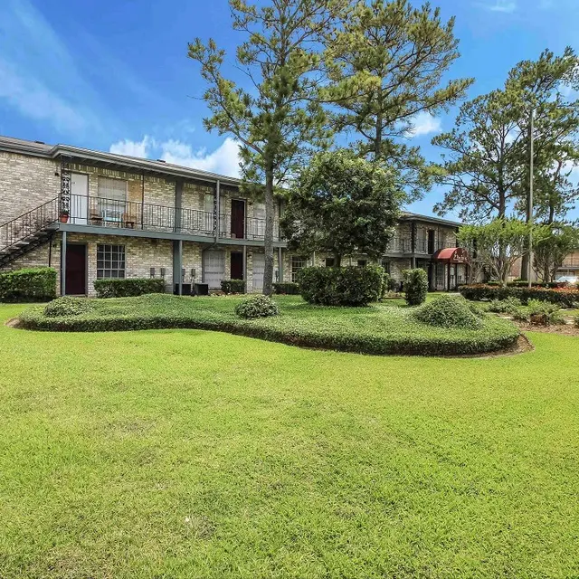 An exterior view of an apartment complex featuring two-story stone buildings surrounded by lush green lawns and trees under a bright blue sky.
