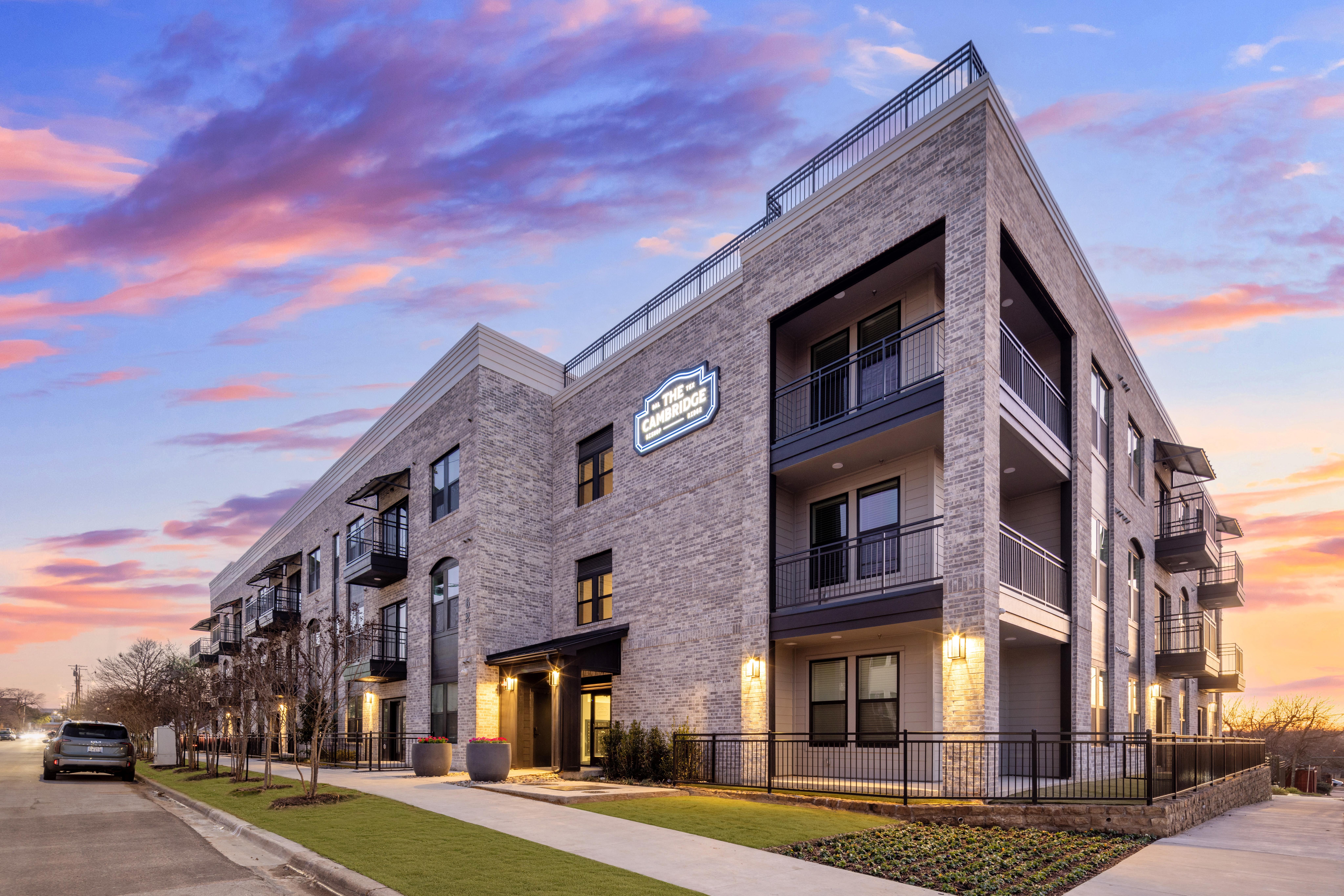 Contemporary Apartment Building at Sunset A modern apartment building with a mix of brick and dark exterior, featuring balconies and outdoor landscaping, set against a colorful sunset sky.