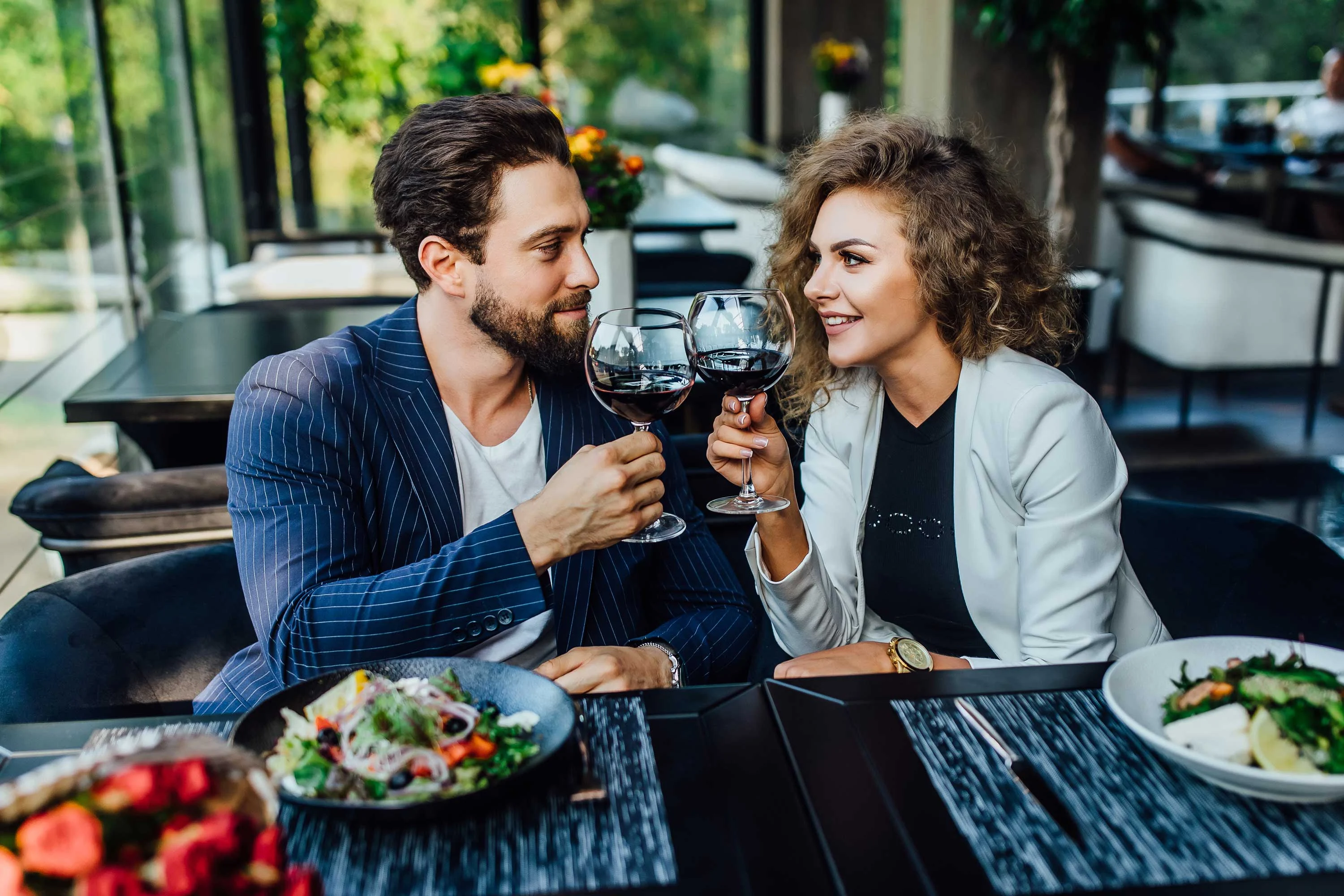 Couple Toasting at Romantic Dinner A couple enjoying a romantic dinner in a restaurant, clinking glasses of red wine. They are seated at a table adorned with plates of salad, surrounded by greenery visible through large windows.