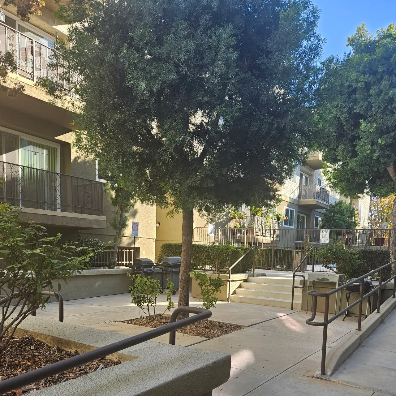 A landscaped walkway in an apartment complex surrounded by greenery and trees, with railings and visible apartments in the background.