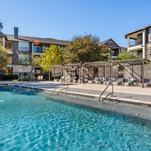 A swimming pool area in an apartment complex featuring a clear blue pool, lounge chairs, and trees in a sunny environment.