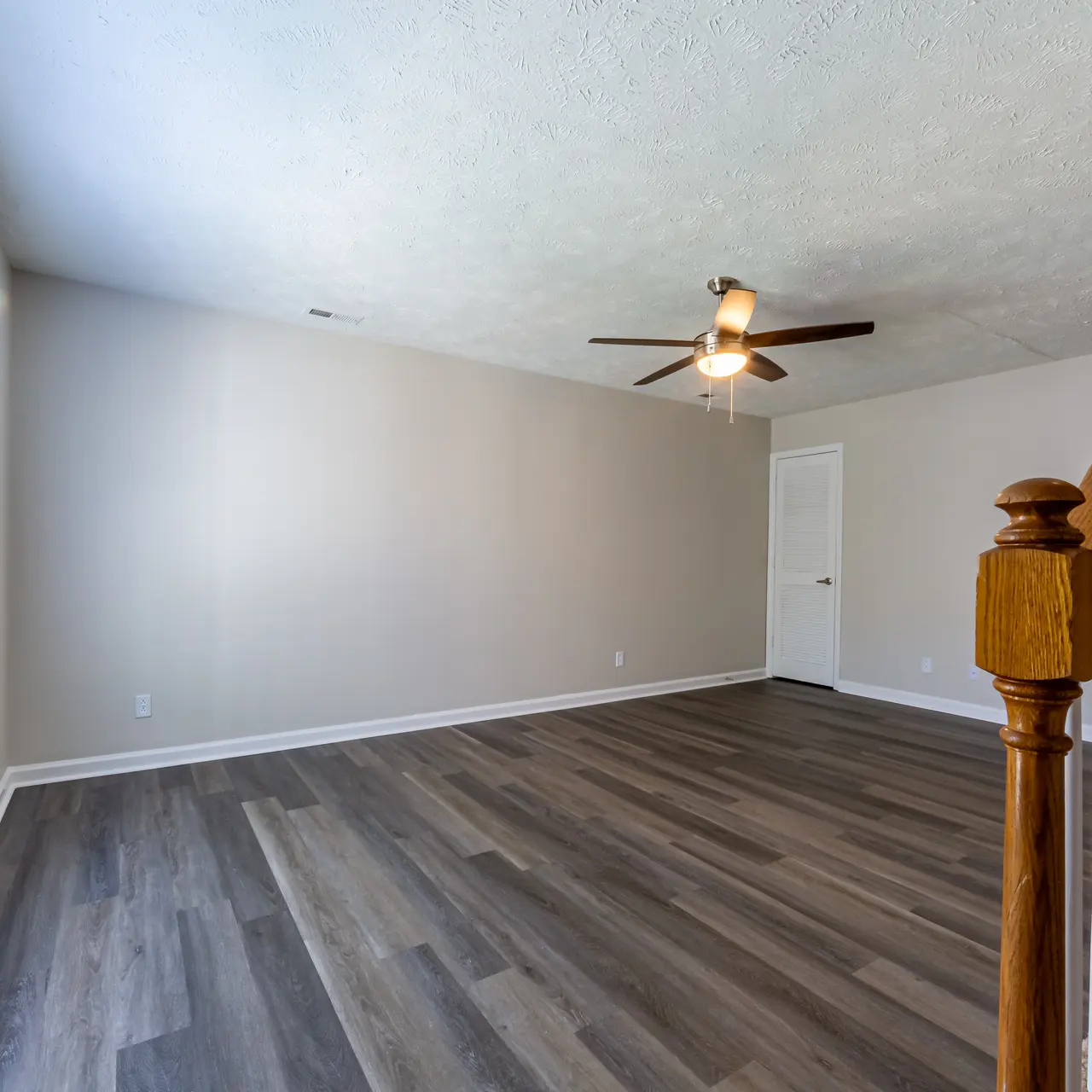 A spacious living room featuring light-colored walls, large windows with blinds, and dark wooden flooring.