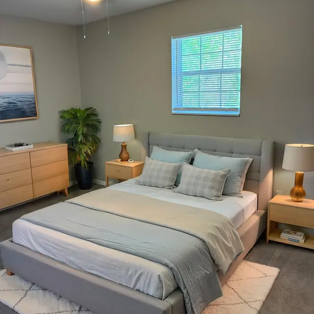 Modern Virtual Staging of a Bedroom A virtual staging of a modern bedroom featuring a gray bed with light bedding, two bedside tables with lamps, a dresser, a window, and a green plant.