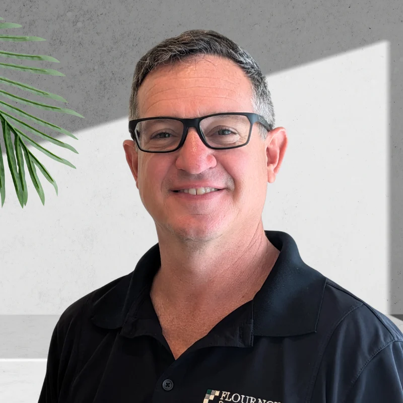 A smiling man wearing glasses and a black shirt with logo, standing in front of a neutral background with leaves from a plant visible on the left.