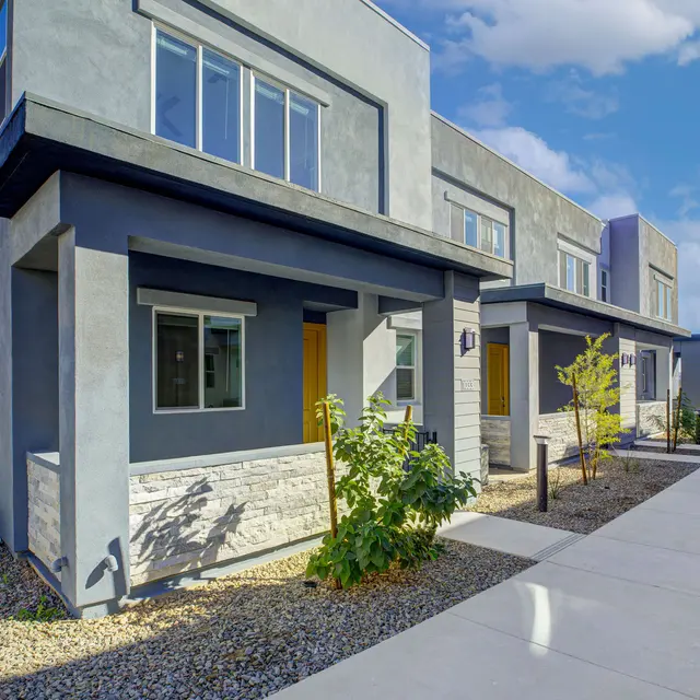 Modern townhouses with gray facades and large windows, surrounded by landscaped pathways and greenery under a blue sky.