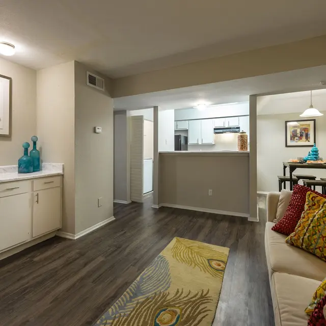 Modern living room with a beige couch, colorful pillows, a decorative rug, and a dining area visible in the background.