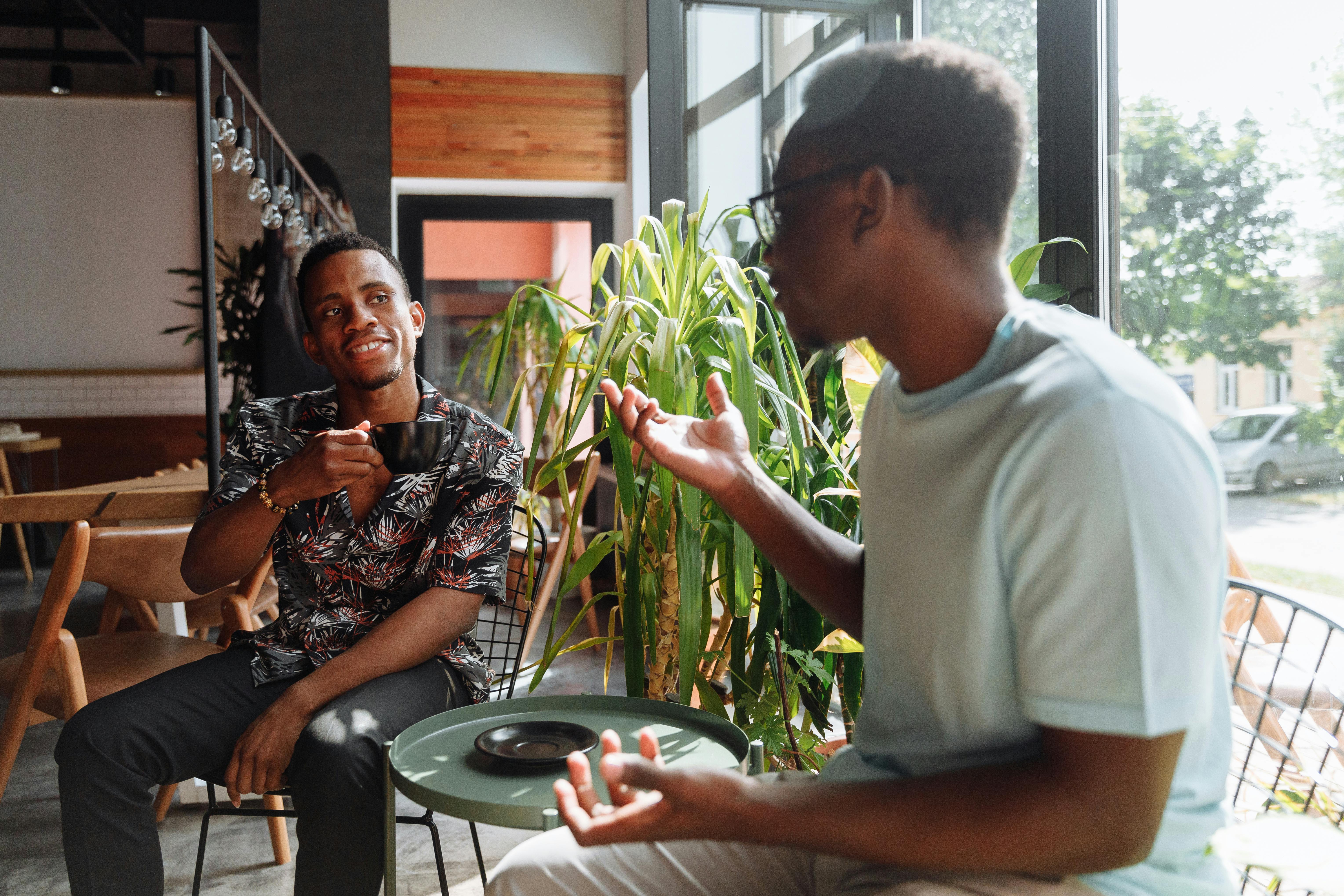 Two men engaged in a lively conversation at a cafe, one holding a cup and smiling, while the other gestures with his hands. The interior features greenery and contemporary decor.